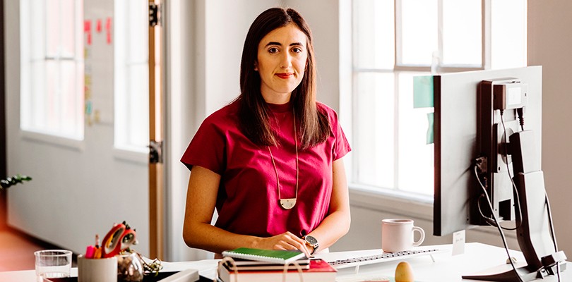 Confident marketing leader standing at her desk, looking at camera. Desk has a a screen, books and coffee mug sitting on it.