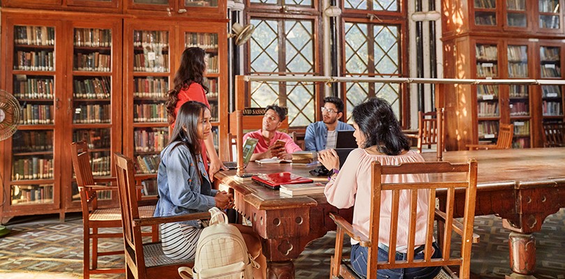 A group of three women and two men, students at a university, sitting around a large desk together in conversation in a library. Books and laptop devices are shown.