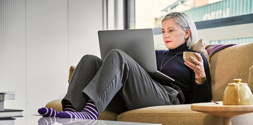 Woman sitting on couch, feet up on table, drinking coffee, looking at a laptop