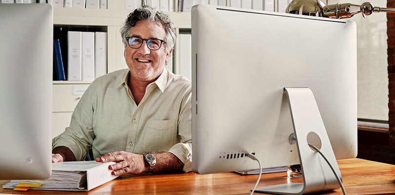 A man smiling sitting at his desk while looking towards the camera. A large bookshelf with design binders lines the background