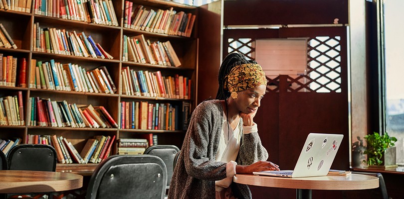 A woman sitting at a small round table in a library with her hand to her chin while working on her laptop.