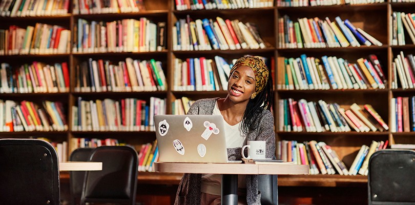 A woman sitting at a small round table in a library with her laptop open while smiling and looking directly at the camera.