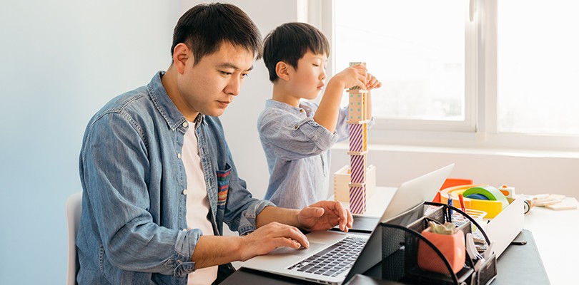 Man working from home on laptop as child plays with blocks nearby