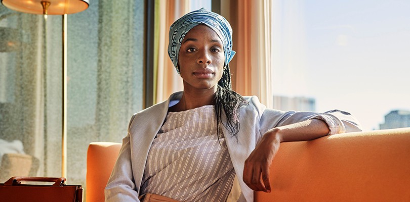 Portrait of a woman looking directly into the camera while sitting down and resting her arm on the back of the couch.