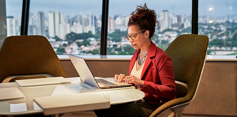 A woman sitting in a chair at a desk working on her laptop in an office overlooking the city.