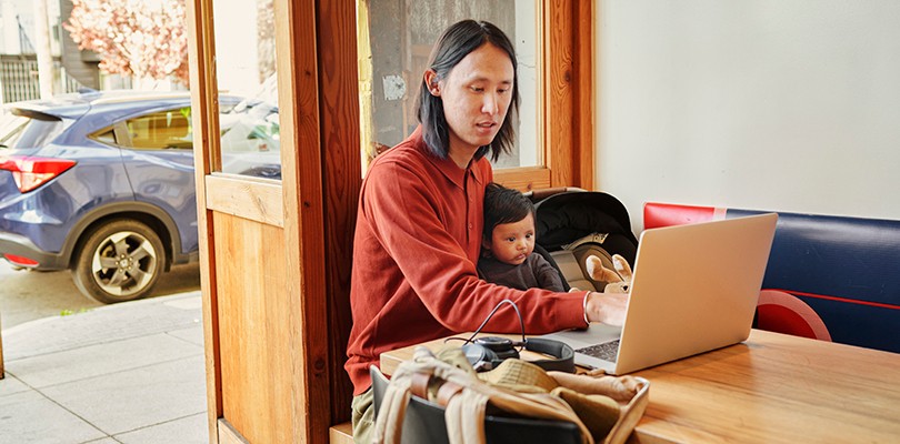 Man working on laptop in a café with baby on lap.