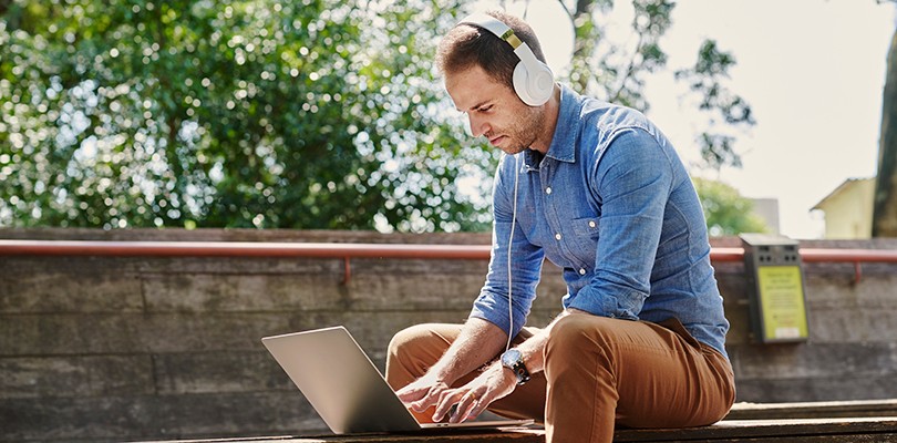 A man sitting outside on a wooden bench with his headphones on while he works on his open laptop.