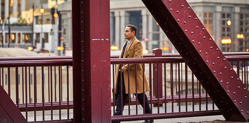 A man in a brown long coat carrying a bag walking across a bridge in downtown Chicago. Large red steel beams from the bridge are shown in the foreground.