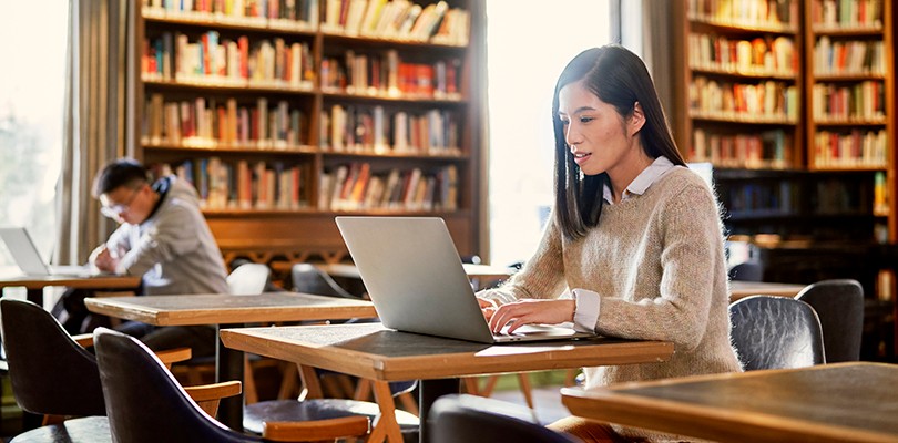 A female student sitting at a library desk focused on her computer. Another male student is seen in the background studying.