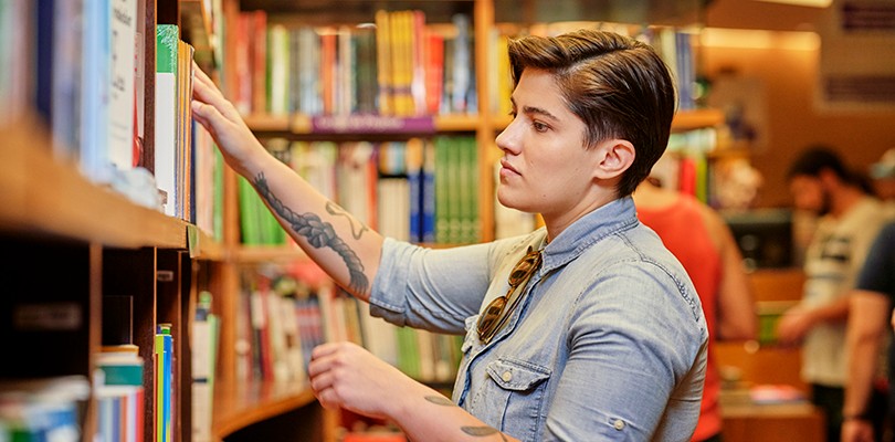 A woman standing inside a library reaching for a book to read, while looking off to a second book.