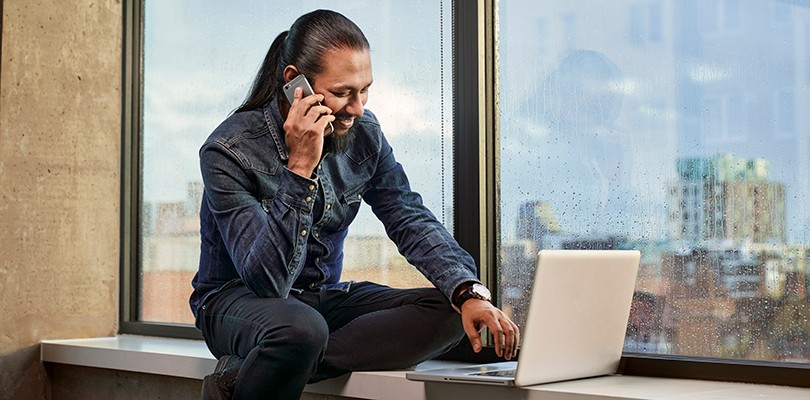 Man on phone sitting in front of window on a rainy day looking at laptop