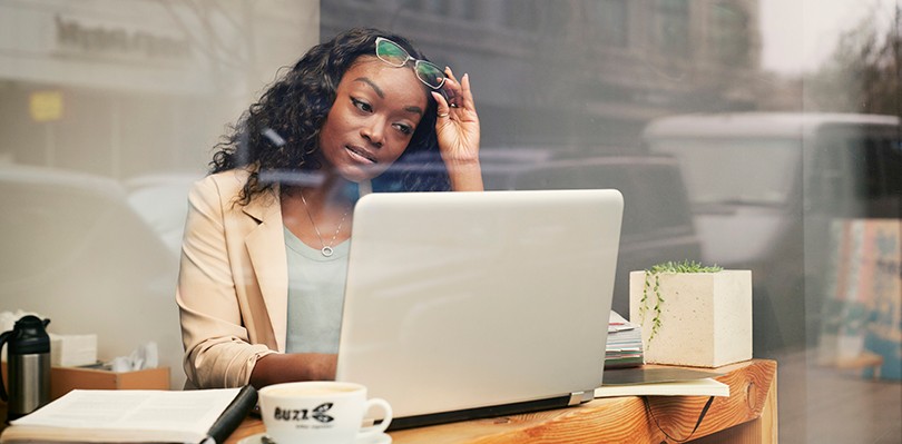 A woman sitting in a cafe counter in front of a window looking out to the street lifts her glasses away from her eyes as she focuses on her computer. The reflection of the exterior is shown in the glass.