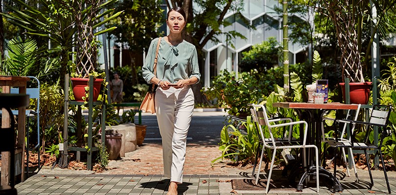 A woman walking outside holding her phone in hand as she looks directly in to the camera. Restaurant tables and lush foliage fill the background.