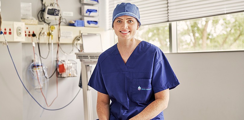 A female nurse is staring at the camera and she is wearing scrubs sitting in a doctor's room. You can see hospital equipment behind her.