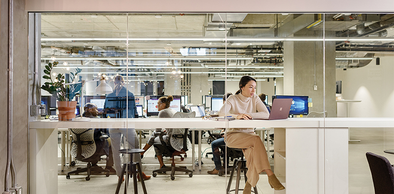 Female office worker seated at high desk in modern office, using Surface Laptop