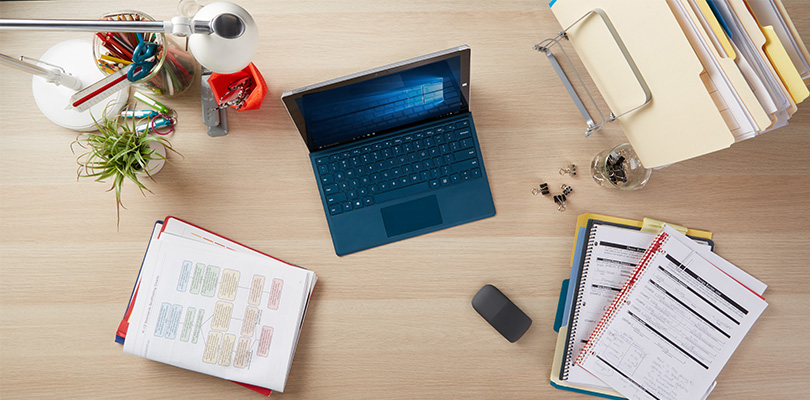 Top of desk view displaying a MIcrosoft Surface with teal keyboard, spiral notebooks, file folders, office supplies, and a plant.