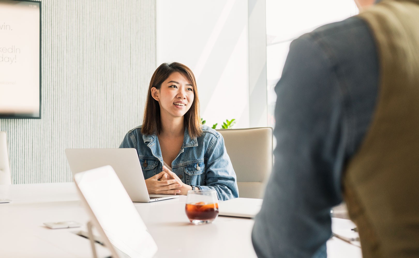 Photo of woman sitting at table with laptop being interviewed by man with back to camera