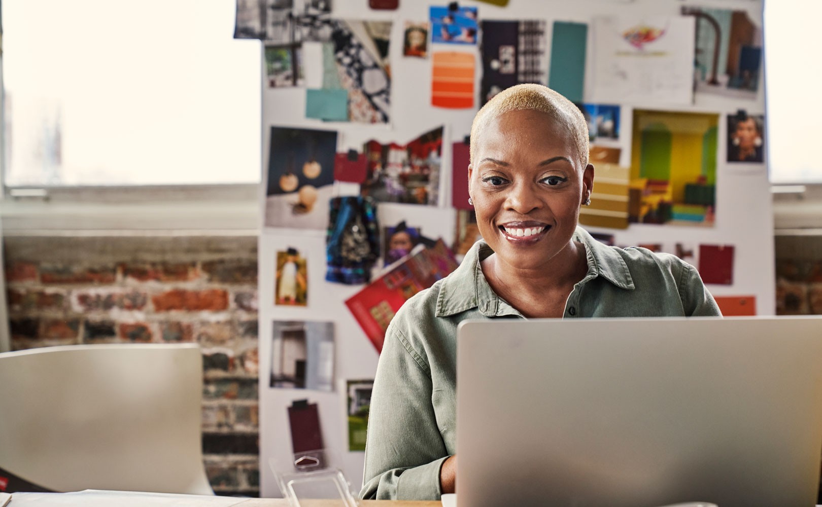 A woman sitting at a desk in an office looking at an image on her laptop that is not pictured. A vision board is displayed in the background.