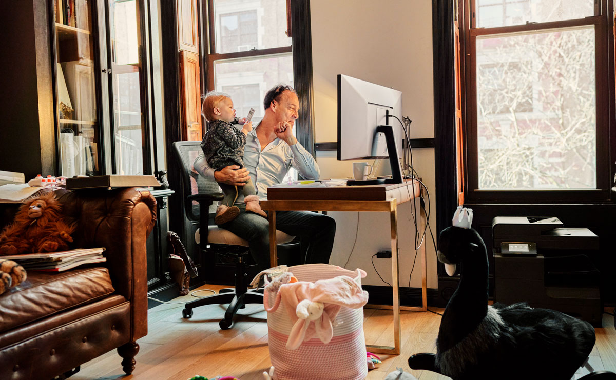 man working from home looking at computer while holding his baby