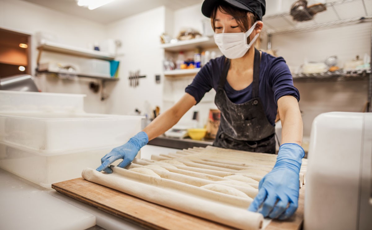 Photo of baker working inside with dough while wearing facemask