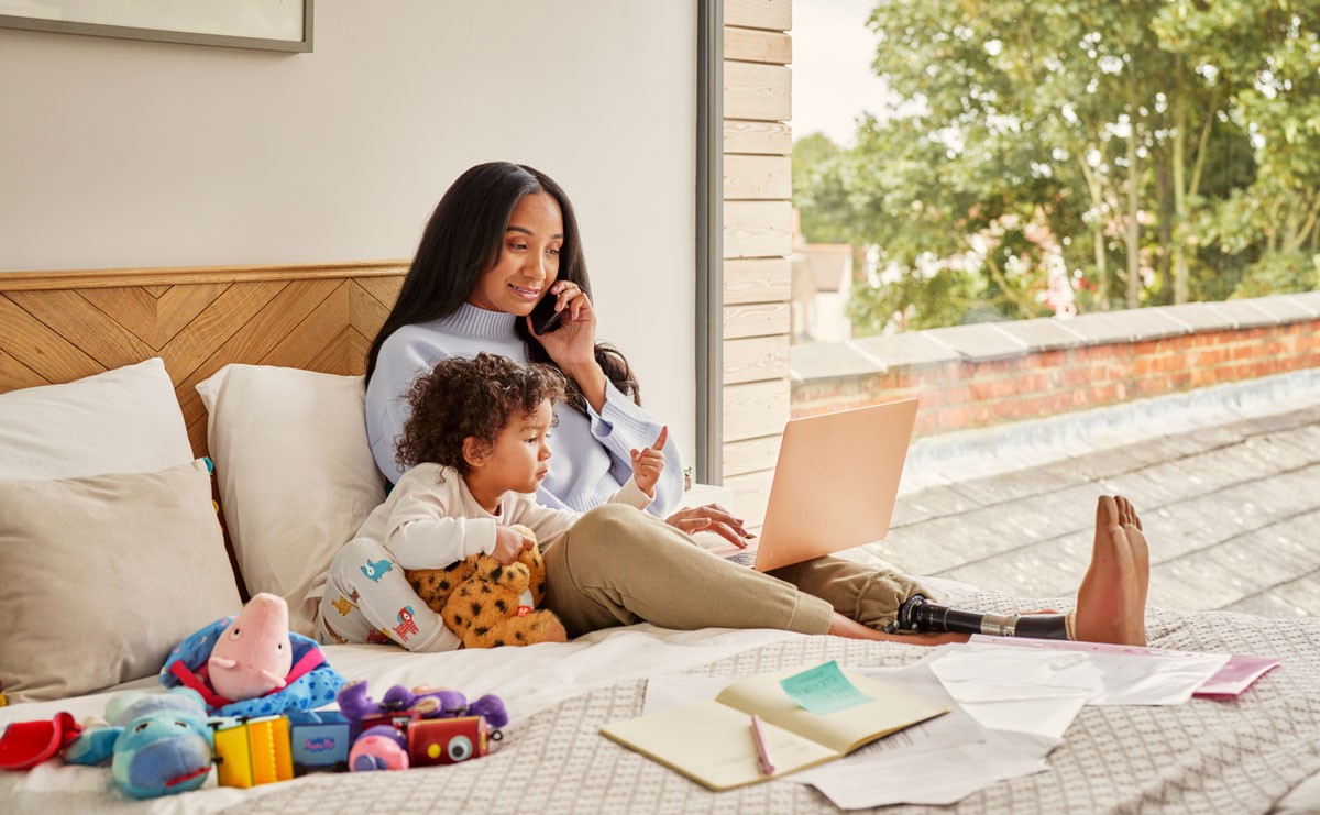 Photograph of woman working from home on bed with child next to her