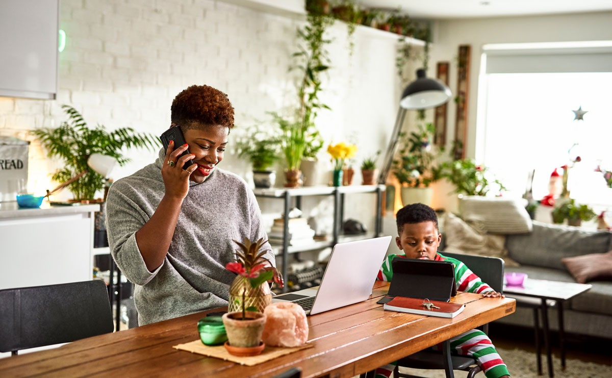 Photo of woman working from home at table with her son playing with a tablet nearby