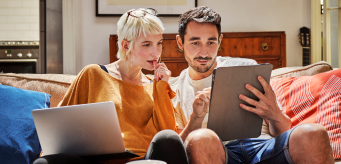 Two individuals sitting down on a couch together and looking at a digital tablet.