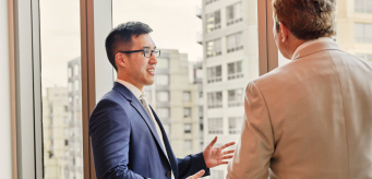 Dois indivíduos conversando e parados ao lado de uma janela.
