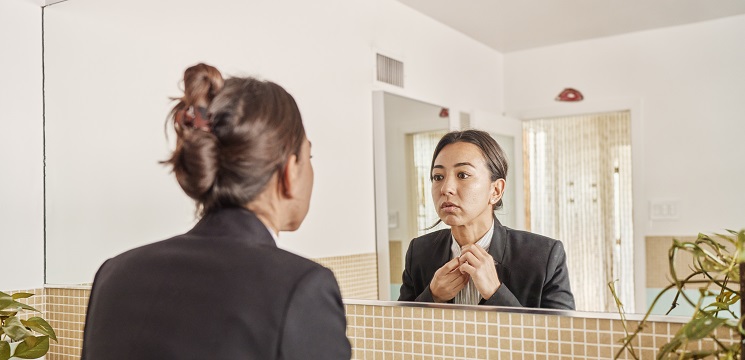 Woman wearing a suit standing in front of a mirror buttoning the top button of her shirt