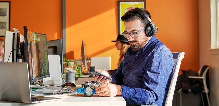 An image of a man in headphones sitting at a computer while looking at his phone.
