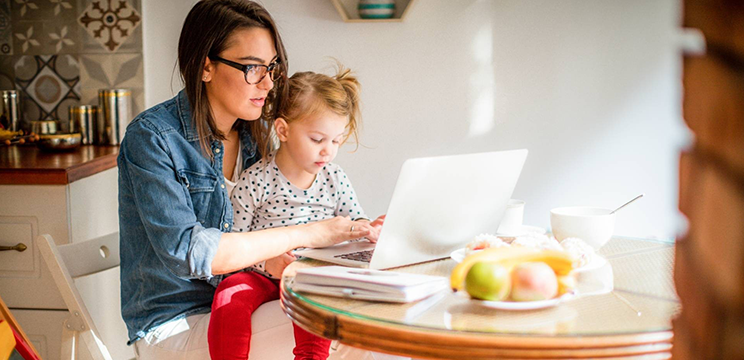 Woman sitting at a table using a laptop with a young girl on her lap. 