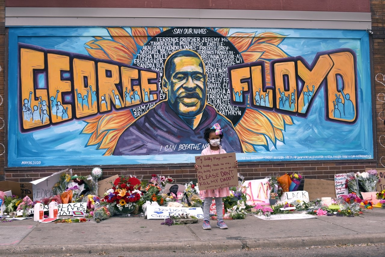 An image of a young girl standing in front of a memorial mural for George Floyd. 