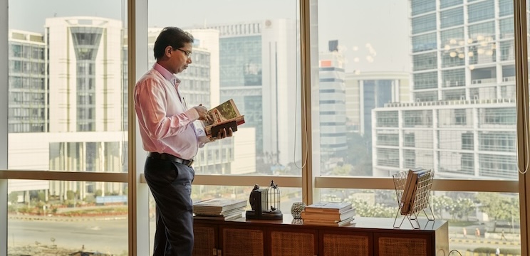 A person is standing by a desk reading a book, looking away from the camera. Sweeping views of Mumbai can be seen out the window.